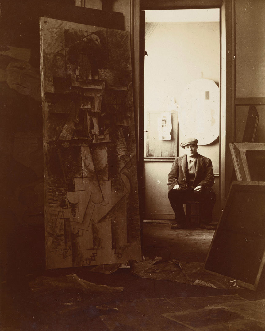 Sepia toned image of a young pablo picasso sitting in his studio with a painting in the foreground. Picasso is framed wearing a hat through the door in the background. 