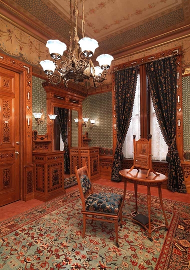 Interior view of the Worsham-Rockefeller room at The Met, with chandelier, embellished wallpaper and ceiling, and detailed woodwork with brass elements.
