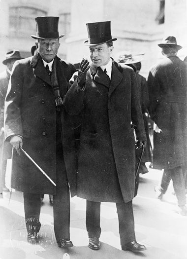 Photo of John D. Rockefeller Sr. (left) and John D. Rockefeller Jr. (right) walking, wearing top hats, suits, gloves, and carrying canes.