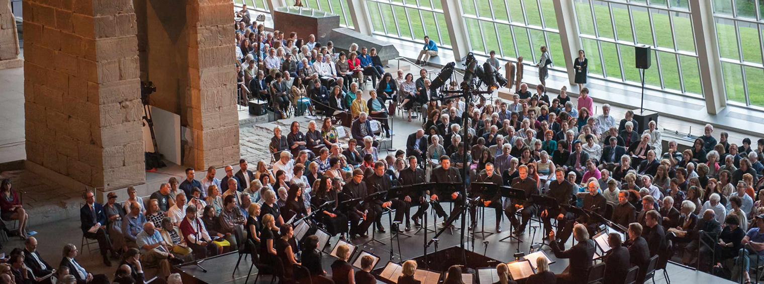 The Estonian Philharmonic Chamber Choir performs Arvo Pärt's Kanon Pokajanen, June 2014. The choir is seated in a circle with spectators seated behind them. The Temple of Dendur is to the left. 