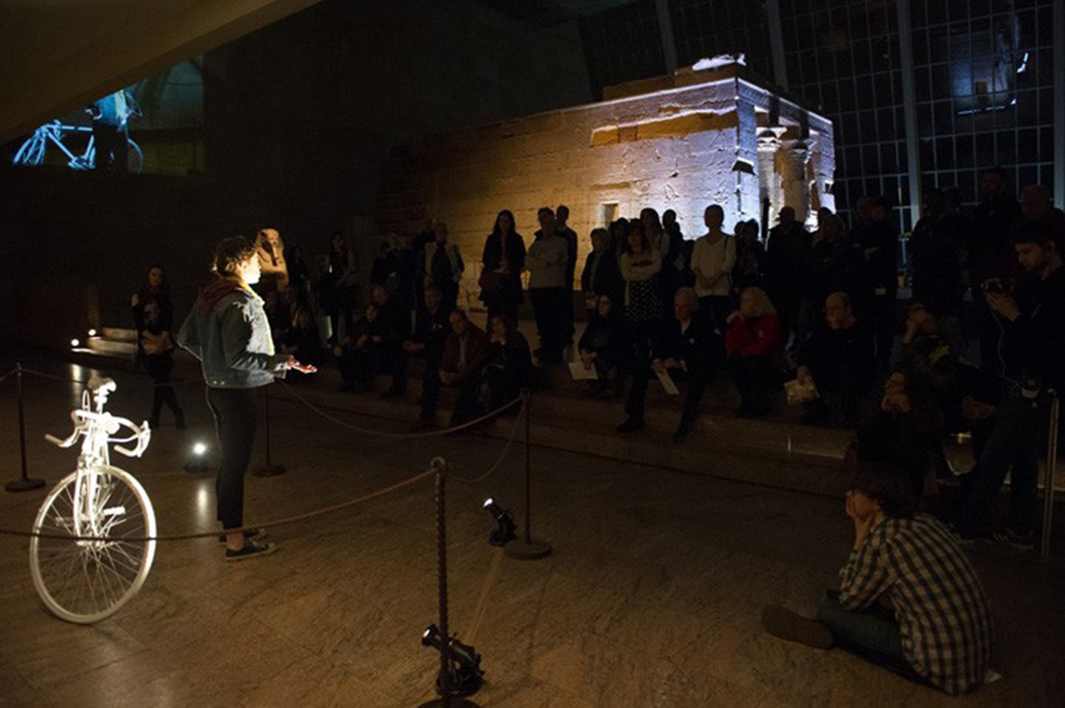 The Civilians, MetLiveArts 2014–15 artist in residence, perform The End and the Beginning outside of the temple, March 2015. A young woman is standing in front of a white bike. The Temple of Dendur is lit up in the background.