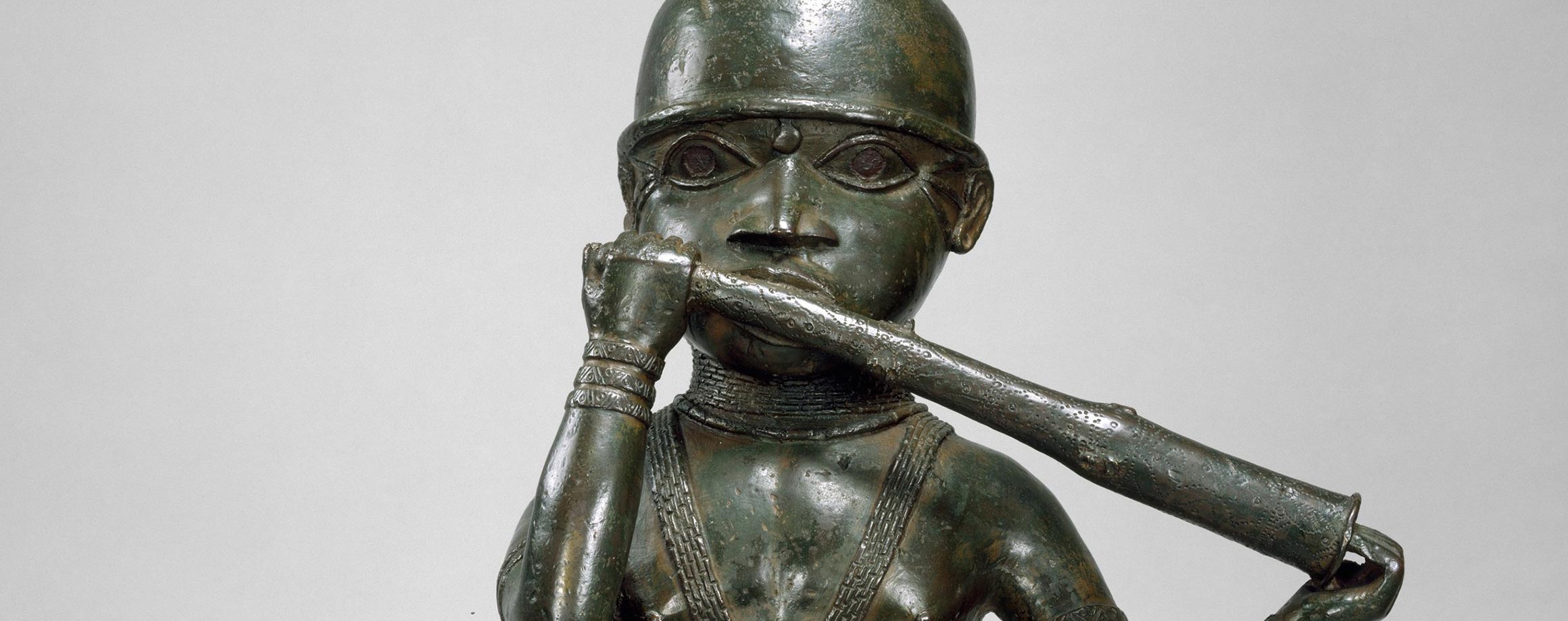 Detail of a Nigerian sculpture, made of dark brass with a green patina and standing two feet tall, of a man playing a horn