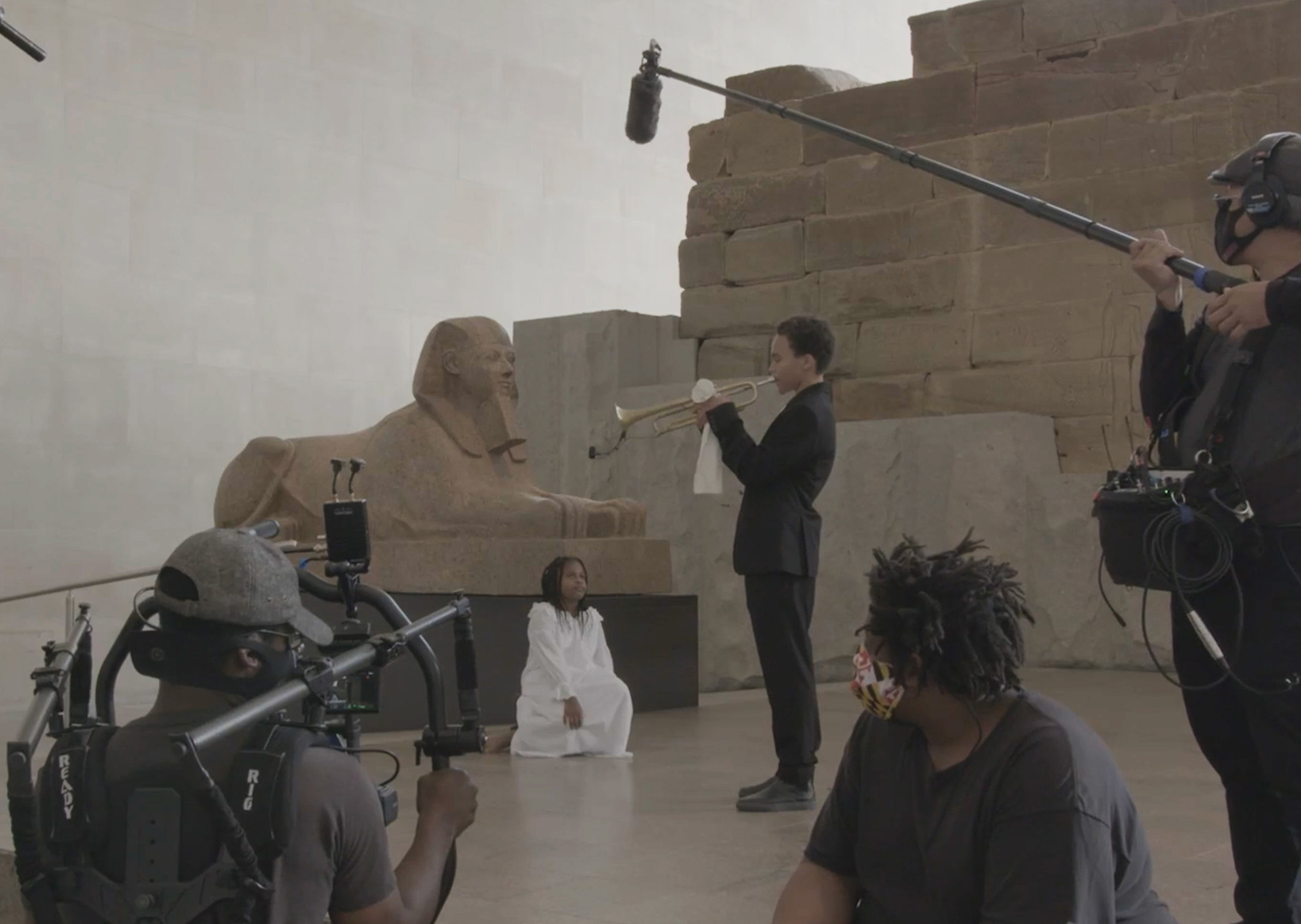 Behind the scenes photograph taken during filming of Rashida Bumbray’s film titled, Untitled, (How High the Moon), 2022; a young girl watches a young man play a trumpet in the Temple of Dendur galleries.