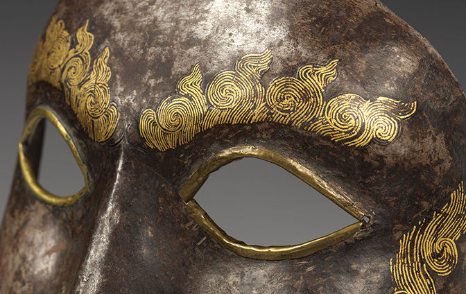 A close up shot of the eyes of a Tibetan War Mask, made of iron that has rusted with gold flame patterns on the eyebrows and wrapped around the eyes. The mask is at a slight angle with a grey background. 