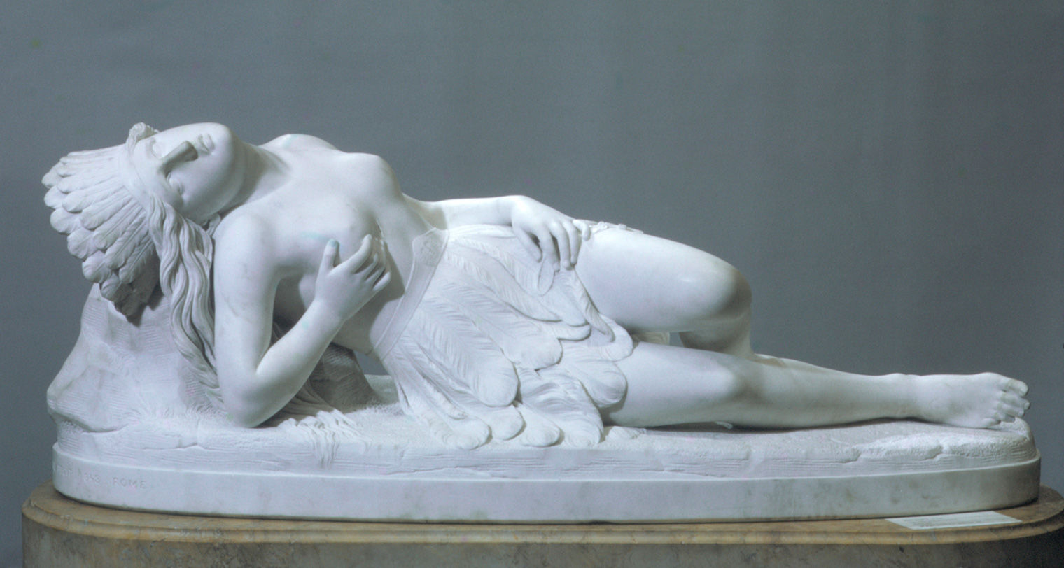 A white marble sculpture of a reclining, bare-chested woman wearing a feathered headdress and skirt, and holding a cross