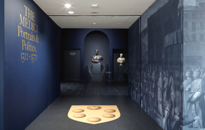 View of the hallway entrance to the exhibition "The Medici: Portraits and Politics, 1512–1570"; the walls are a deep sapphire blue and the title and the design of the Medici family's coat of arms on the floor are both a warm gold color; a sculptural portrait bust appears within a niche at the end of the hall entrance