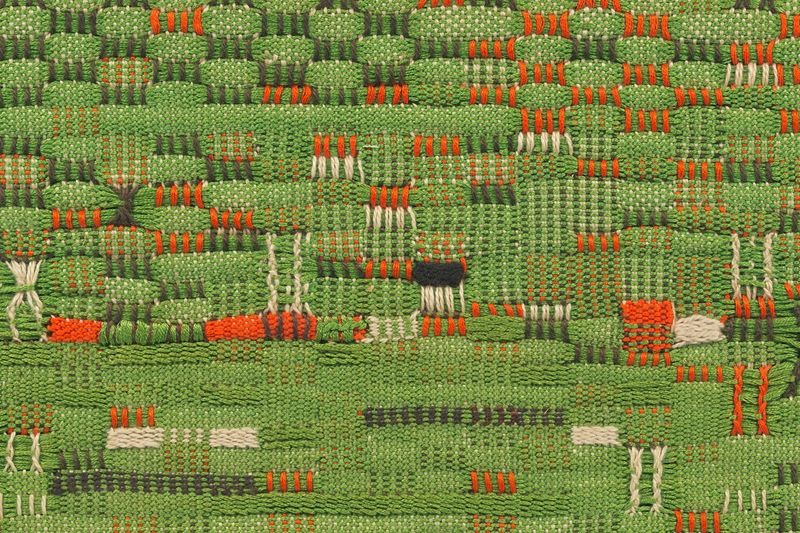 Detail of a woven material with grass green as the primary color, accented with dark green, oranges, and whites.