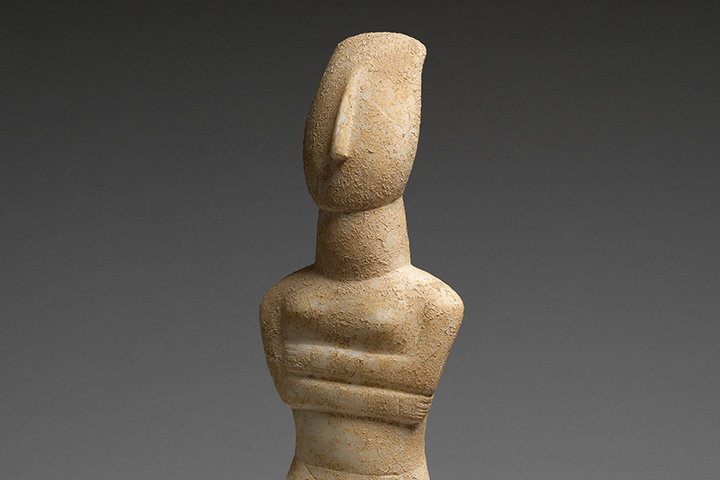 Nude stone Cycladic figure with its arms crossed against a gray background. 