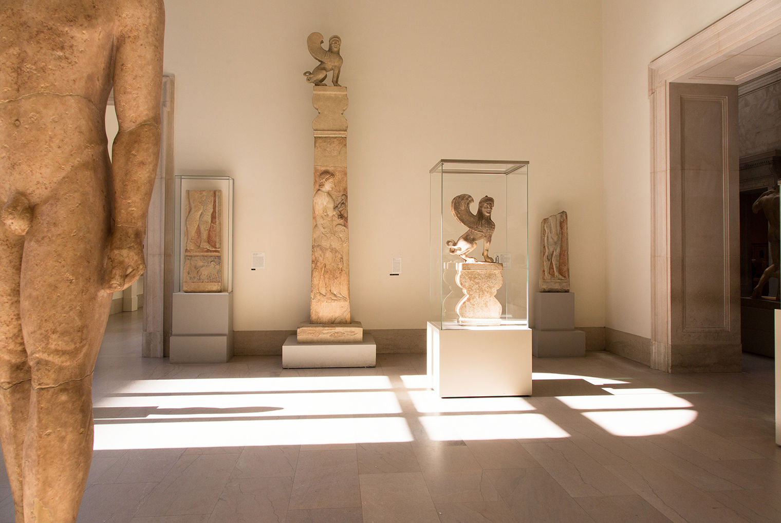 A sunlit Gallery 154 at The Met. Beyond a nude male sculpture in the left foreground, the tall stele is located along the back wall, between two marble stone fragments. The crowning sphinx is in a glass case in the center of the room.