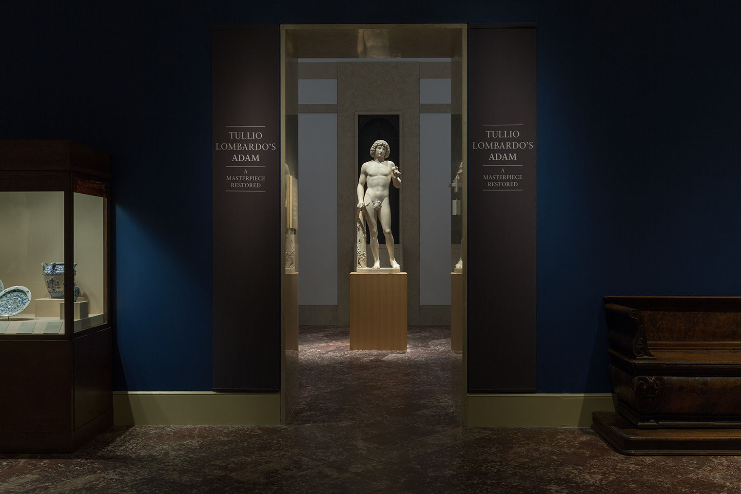 Light is trained on Adam, standing on a wood pedestal and centered within a doorway. Signage around the doorframe announces the exhibit celebrating his restoration.
